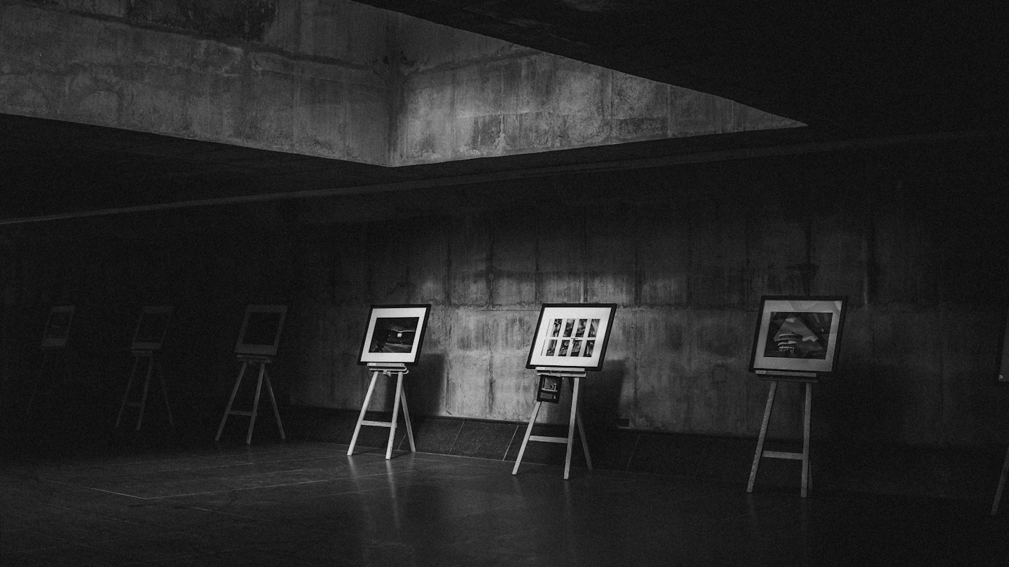 Framed artworks stand on wooden easels in a concrete gallery.